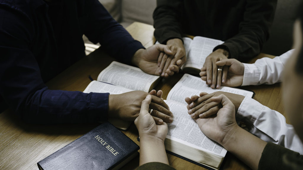 A diverse group of Christians holds hands while praying during Bible study showing the strength in unity.