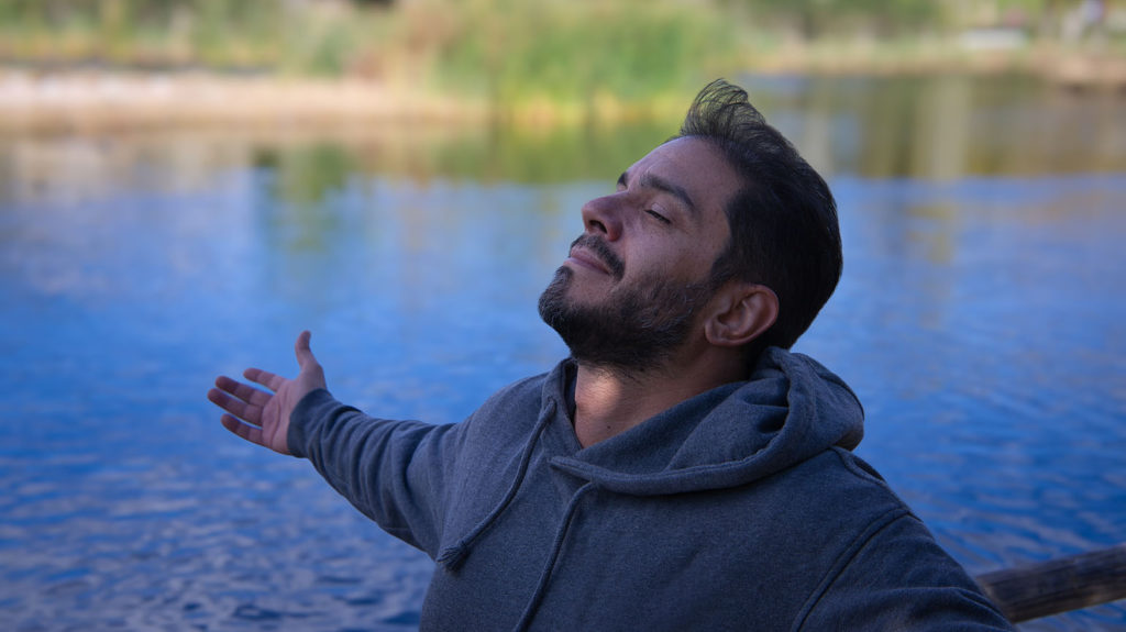 A young man in a blue hoodie stands beside a lake with open arms enjoying God's goodness.