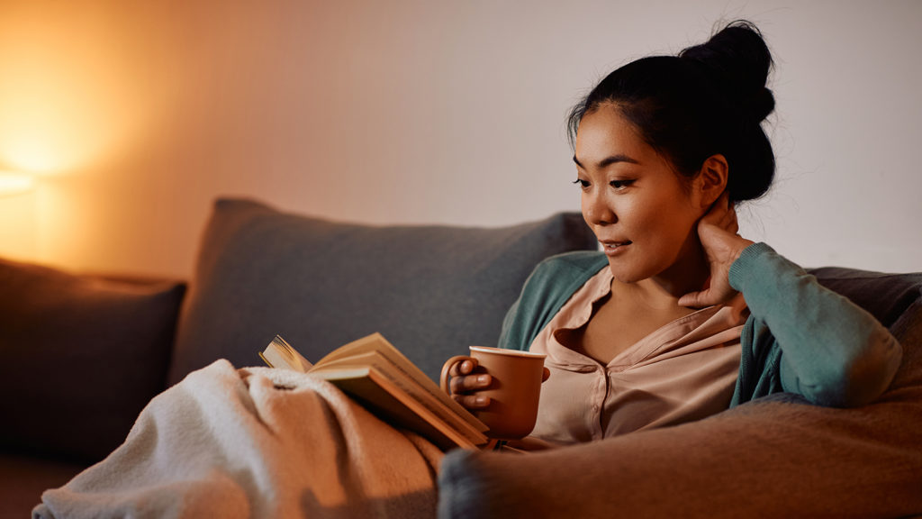 Young woman drinking tea on the couch while she reads so she sleeps better