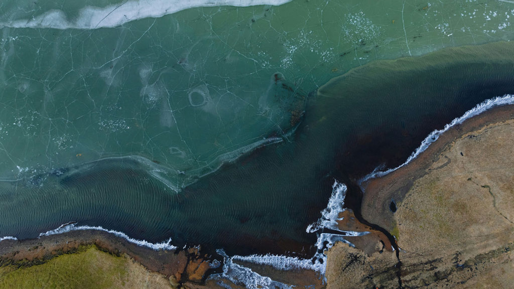 An aerial shot of ocean waves touching the bottom of a cliff remind us that we are cared for by God all the time.