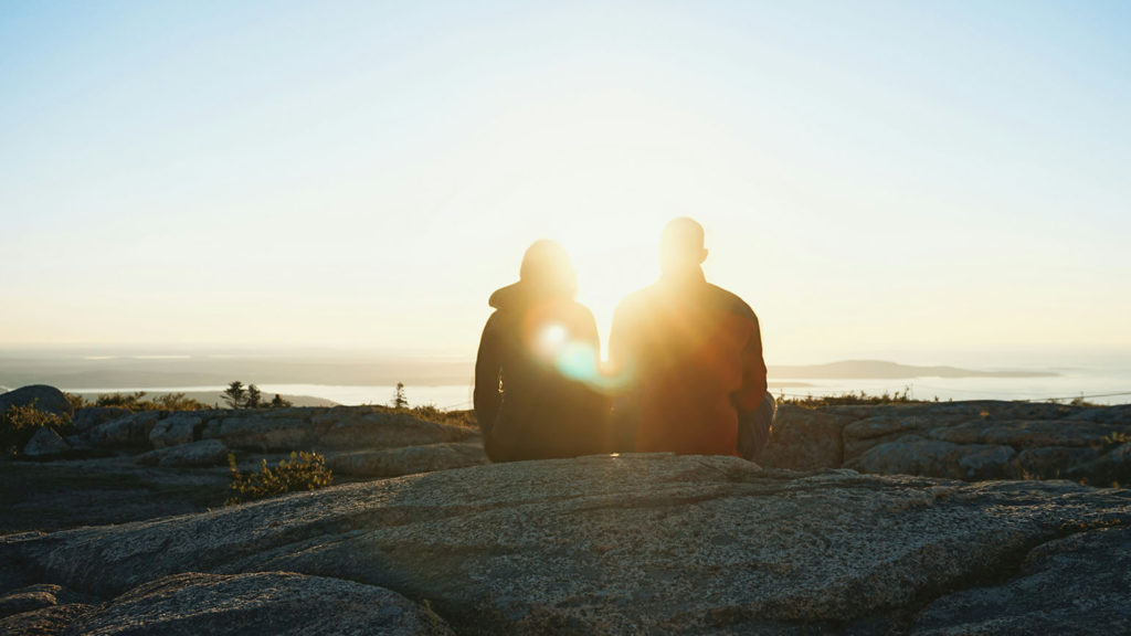 Two people sit overlooking the ocean at sunset practicing healthy conflict resolution.