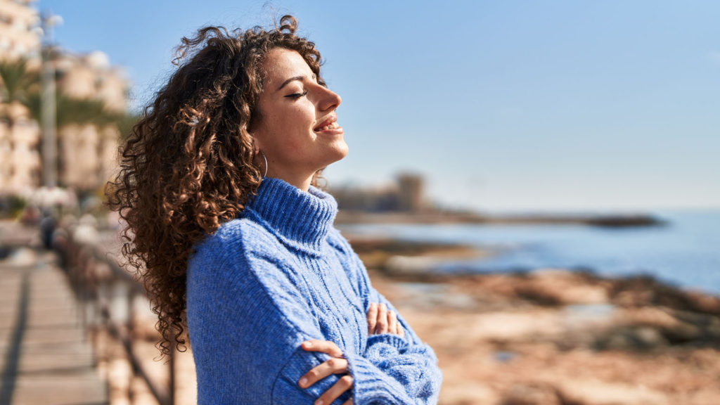 A young woman in a blue turtleneck sweater can cultivate peace outdoors by the seashore.