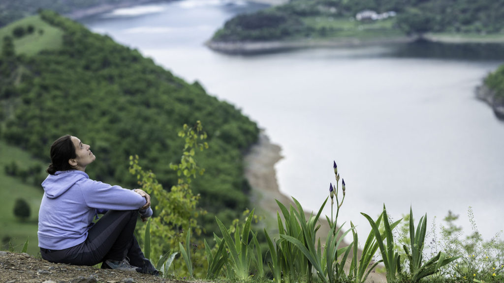 A young woman in a blue hoodie is experiencing God with all her sense on a mountain by a lake.