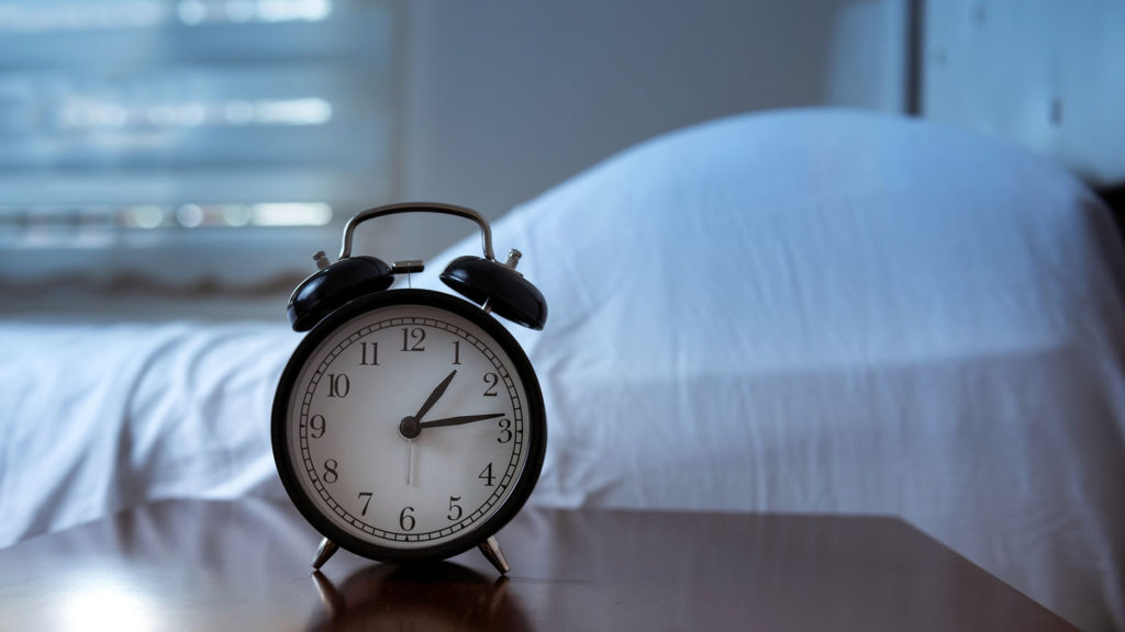 An alarm clock showing 1:15 a.m. sits on a table beside an empty bed showing that it's owner is not getting enough sleep.
