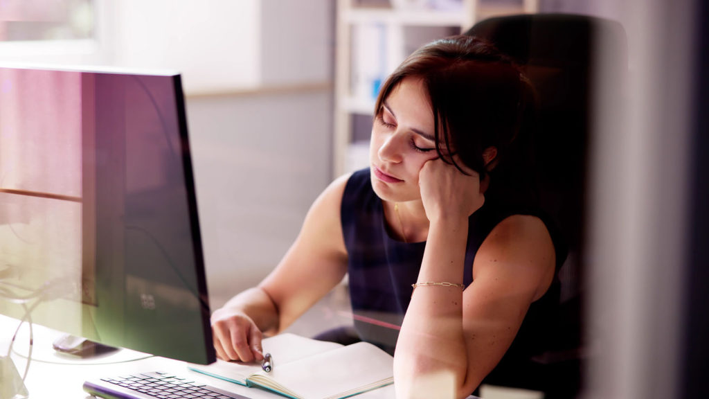 A woman in a black dress sits at her computer with her eyes closed and her head resting on her fist because she's not getting enough sleep.