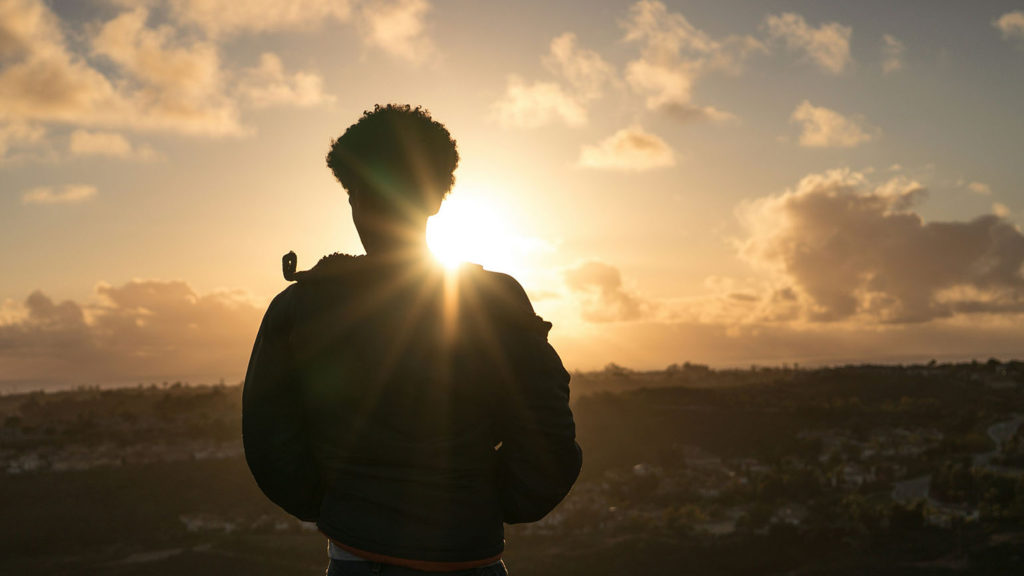 A silhouetted young man stands before a sunset knowing he needs to return to God.