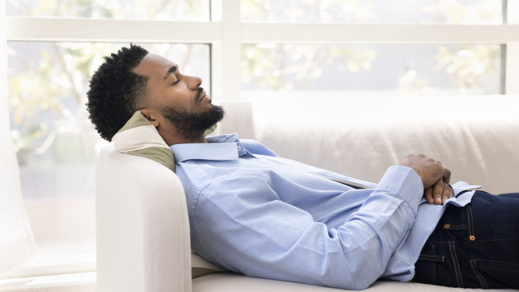 A young Black man takes a nap on a white couch as he does shift work and sleep isn't always easy.