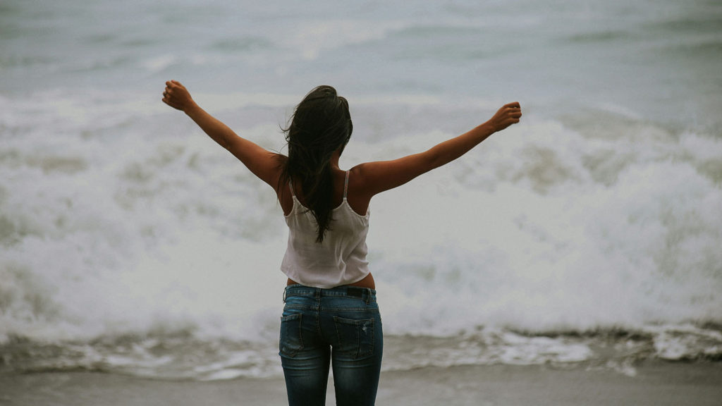 A young woman stands on the shore with arms upraised as she found the key between sleep and energy.