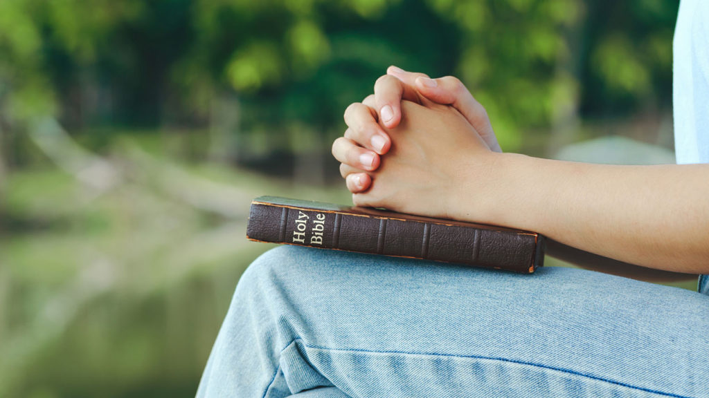 A woman sits outside with her hands folded on a closed Bible as she enjoys the spiritual benefits of sleep.