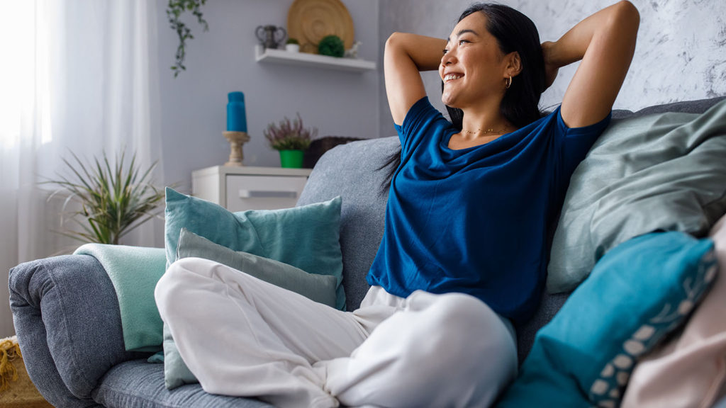 A young Asian woman sits on a couch with her hands behind her head and a smile on her face as she discovered the spiritual weapon of good sleep.