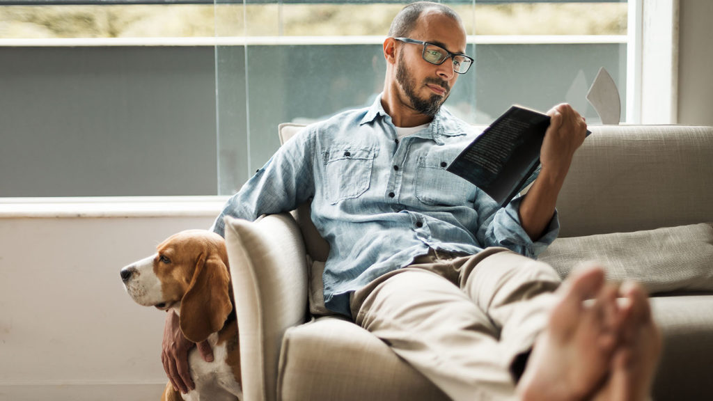 A bearded man with glasses sits on the couch petting his pet beagle while reading his Bible to discover the spiritual weapon sleep brings.