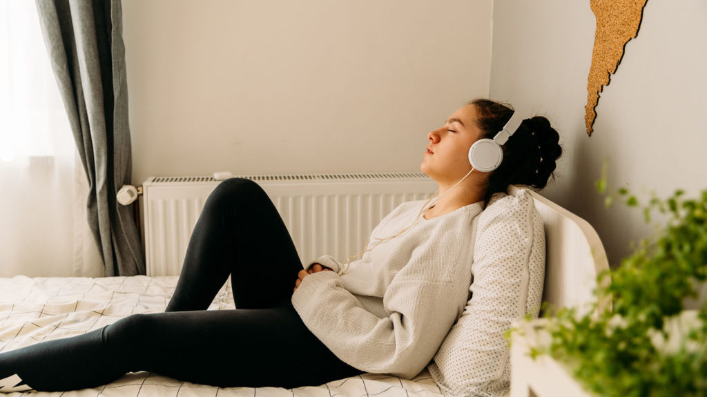 A young woman reclines on her bed with headphones listening to soothing music as she is winding down before bedtime.