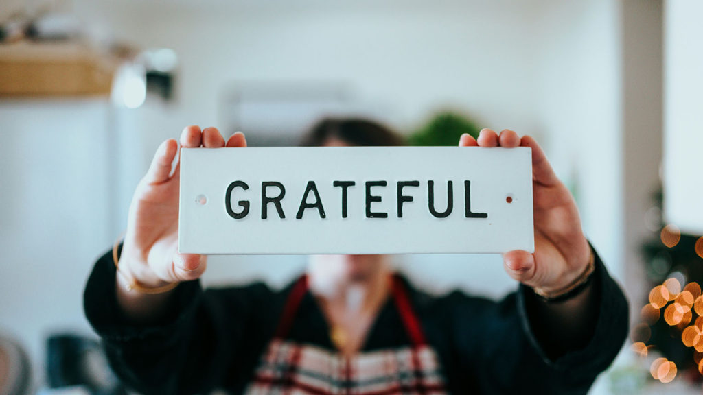 A person holds up a small wooden sign saying "grateful" as they consider the gratitude meaning they adopt.