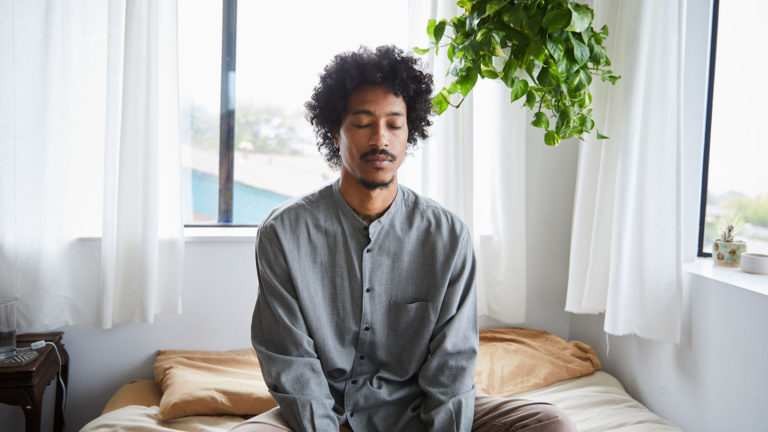 Young African American man sitting with his legs crossed and eyes closed on his bed as he recites prayer Bible verses to start his day.