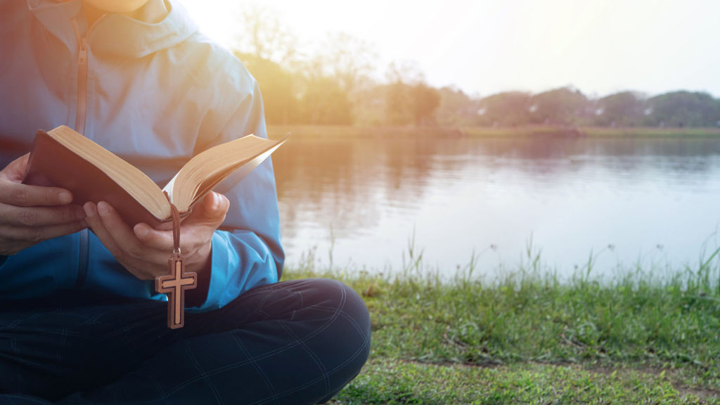 A man reads his Bible by a lake in the morning looking for prayer Bible verses to start his day.