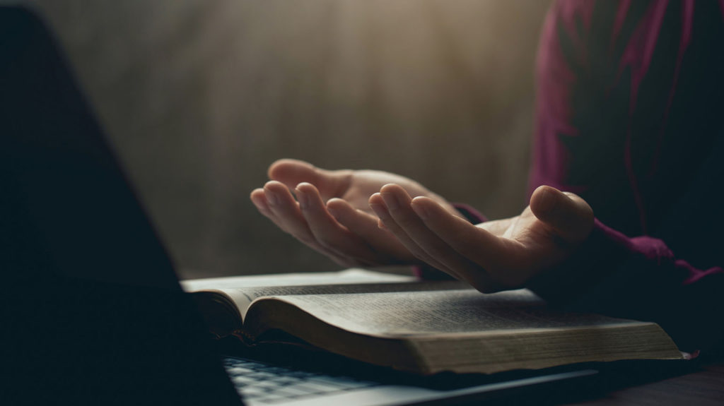 A woman holds her hands palm up over an open Bible as she prays a prayer Bible verse for God's will to be done.