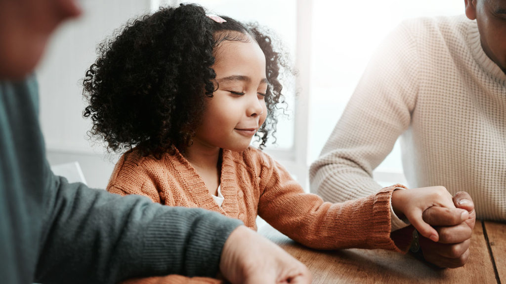 A young black girl holds hands with her family as she prays with them over their prayer board.