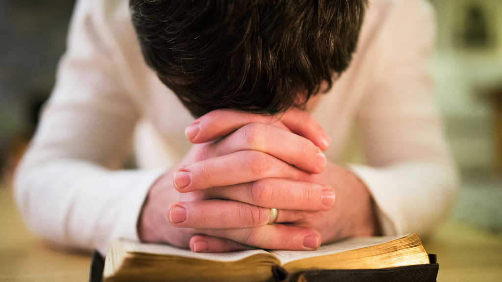 A woman has her head bowed and hands clasped over an open Bible as she prays a prayer for today.