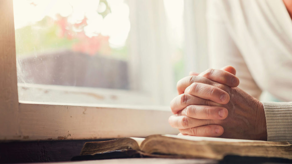 Near a bright window, I woman sits with her hands folded in prayer on her open Bible as she combines prayer with Scripture each morning.