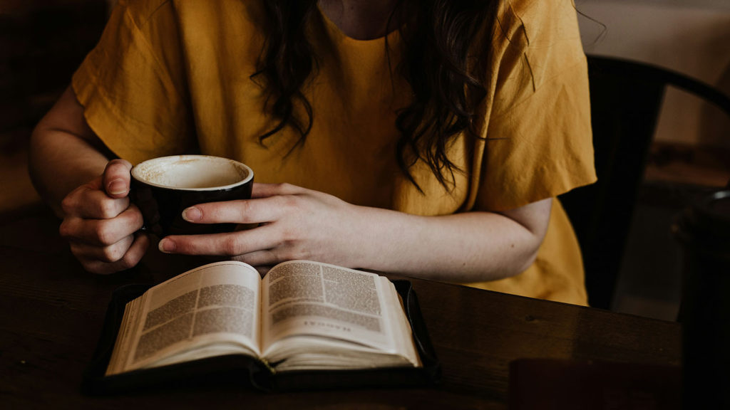 A woman in an orange blouse holds a cup of coffee as she reads her Bible in order to combine prayer with Scripture to hear from God.